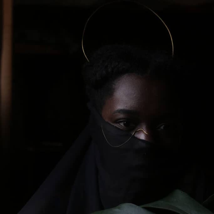 Image Description: A portrait of a dark-skinned African woman’s face obscured by a black transparent veil that runs just under her eyes covering the bottom of her face. Visible under the veil is gold jewelry wire running over her nose, curving under her c