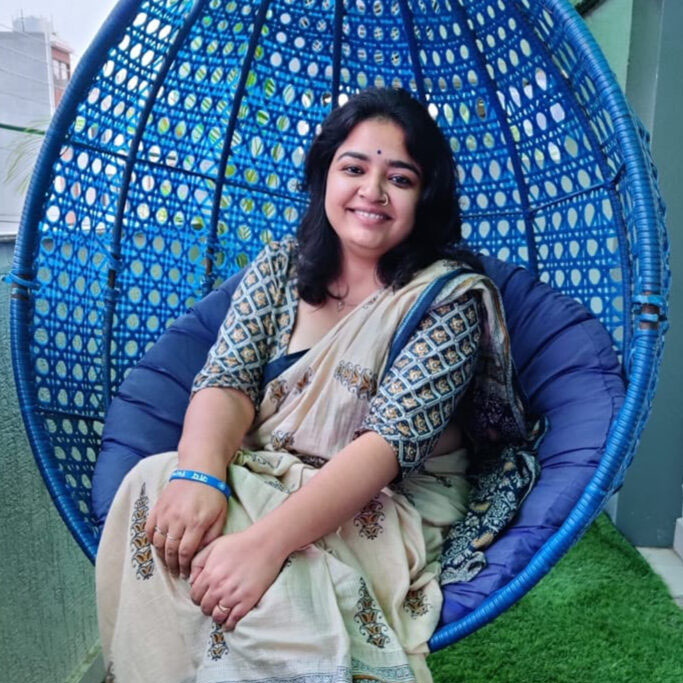 A femme presenting person is sitting in the balcony of room on a blue couch swing. She is wearing a cream colour saree with dark blue border and blue printed blouse. She is also wearing a bindi, a nose ring and a blue band on her left hand that says Jai B